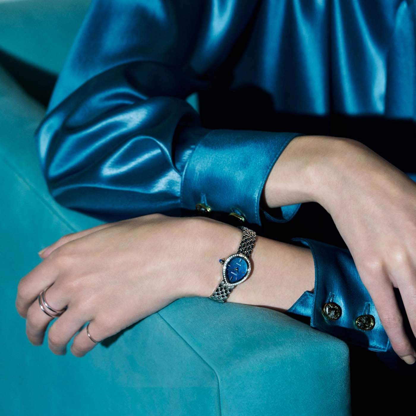 A woman in a blue satin top and rings wears a watch.