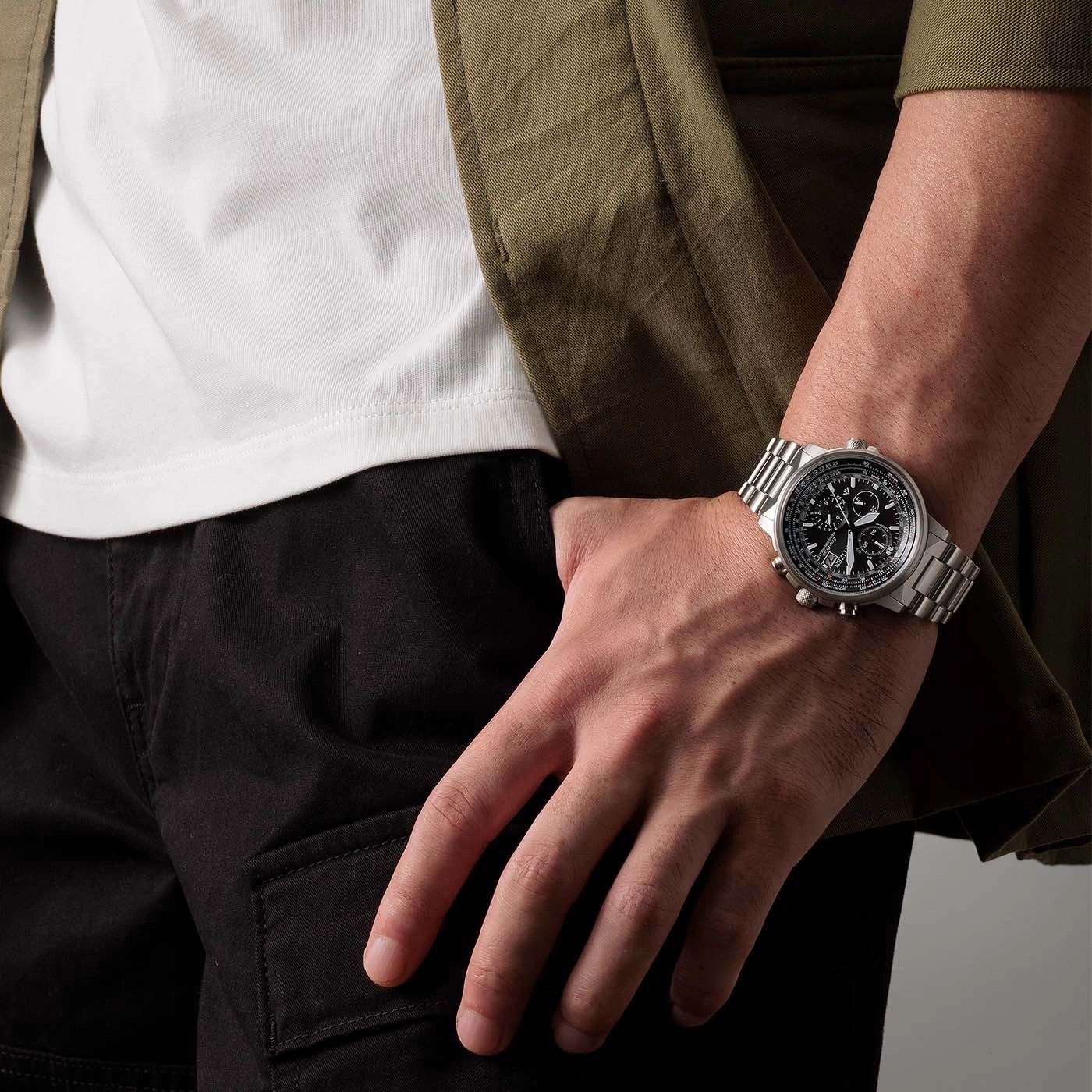 Man wearing a watch, black pants, white shirt, and green jacket.