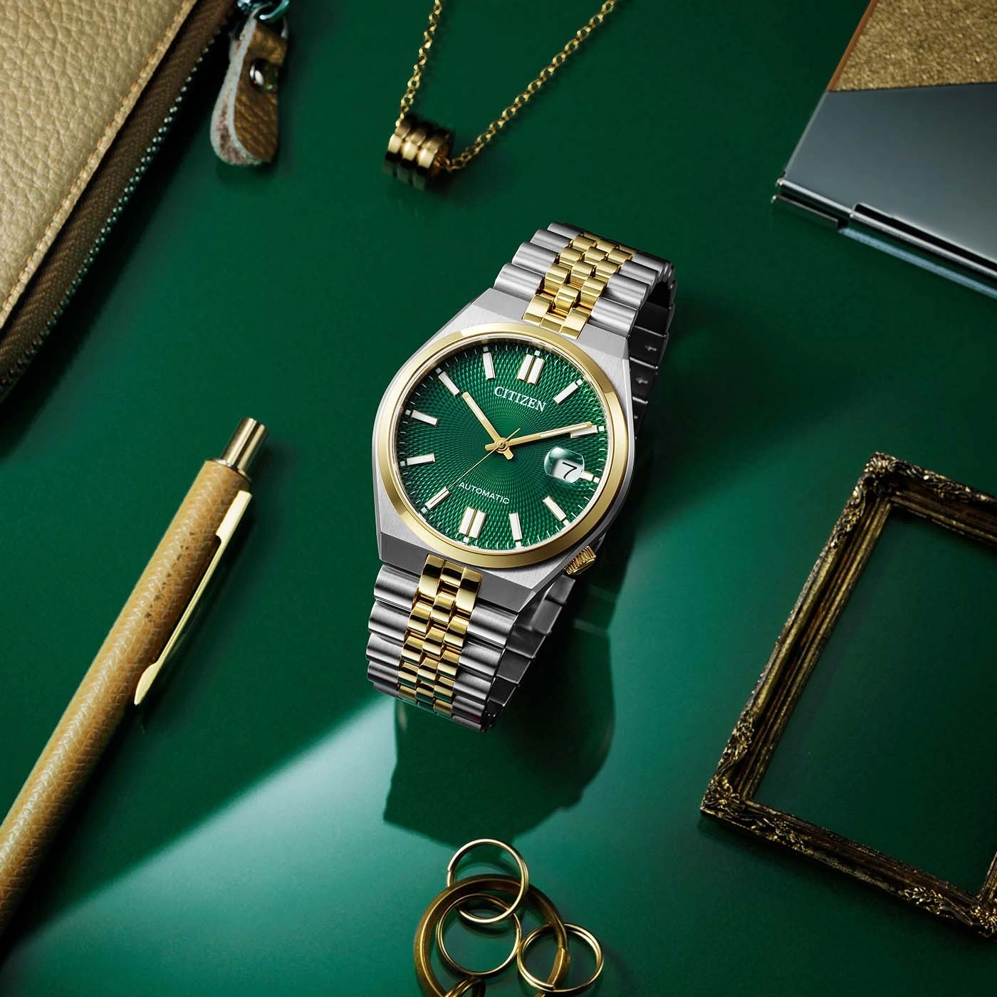 Citizen watch with green face, gold and silver bracelet, on green surface with accessories.