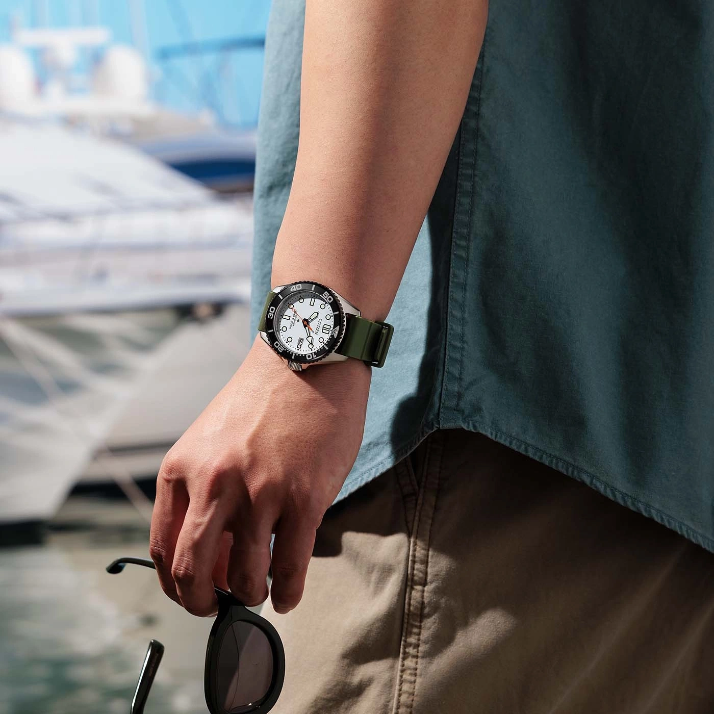 Man wearing a watch and holding sunglasses by the water with boats in the background.