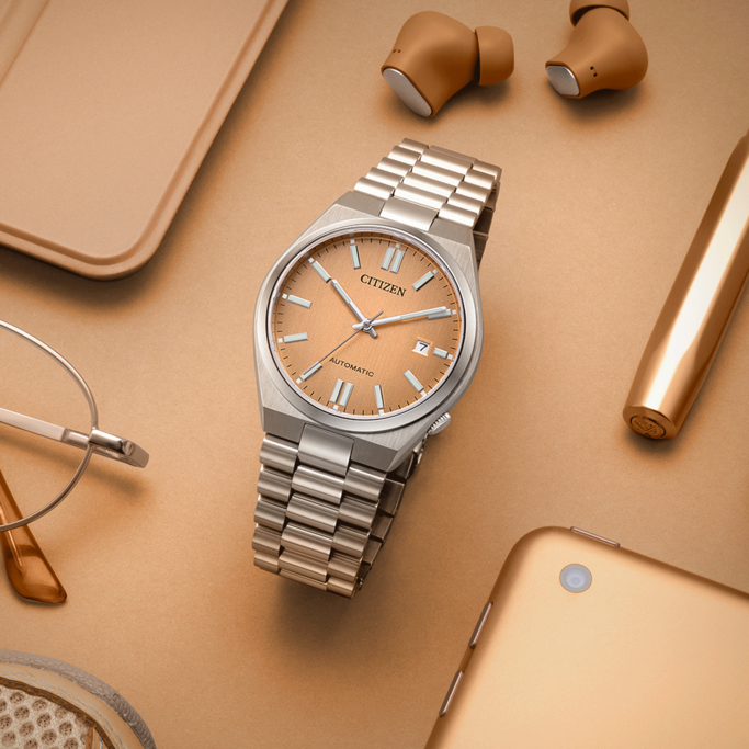 Citizen watch, earbuds, pen, glasses, and a tablet on a gold surface.