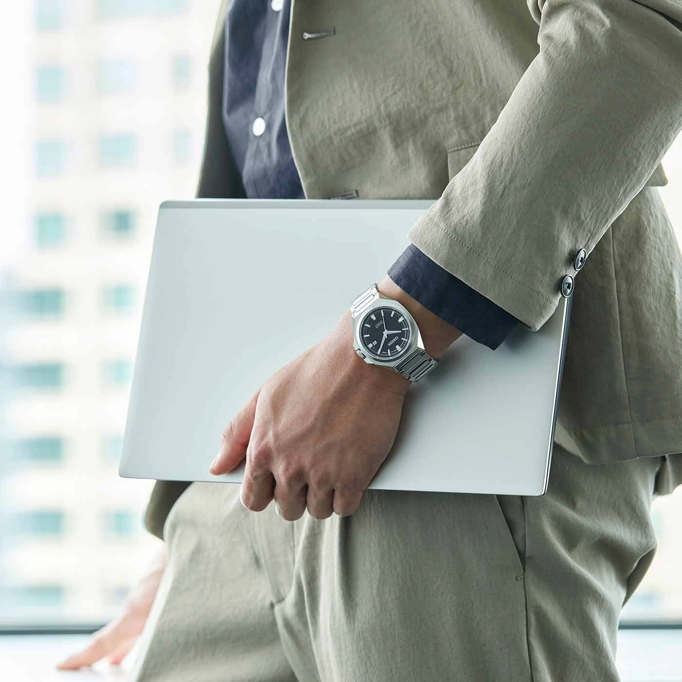 Person holding a laptop, wearing a watch and a suit, near a window.