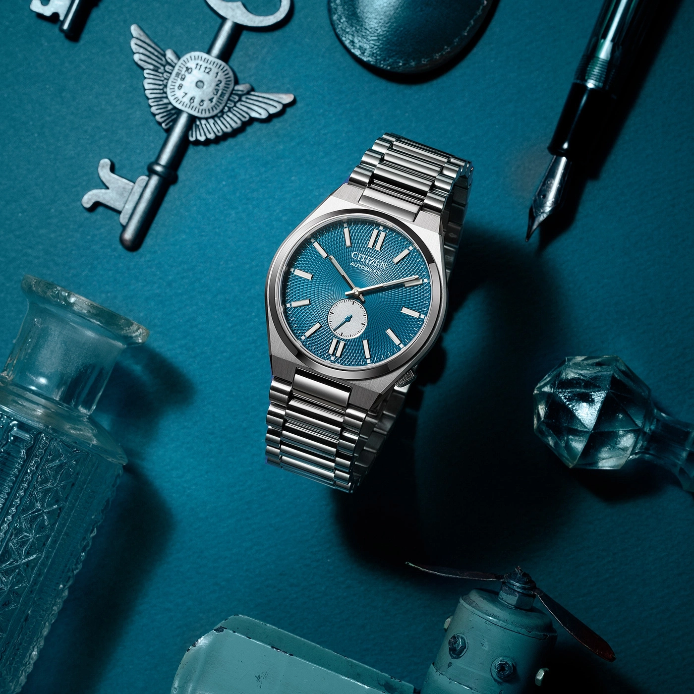 Citizen watch with blue face, keys, pen, and other objects on blue surface.