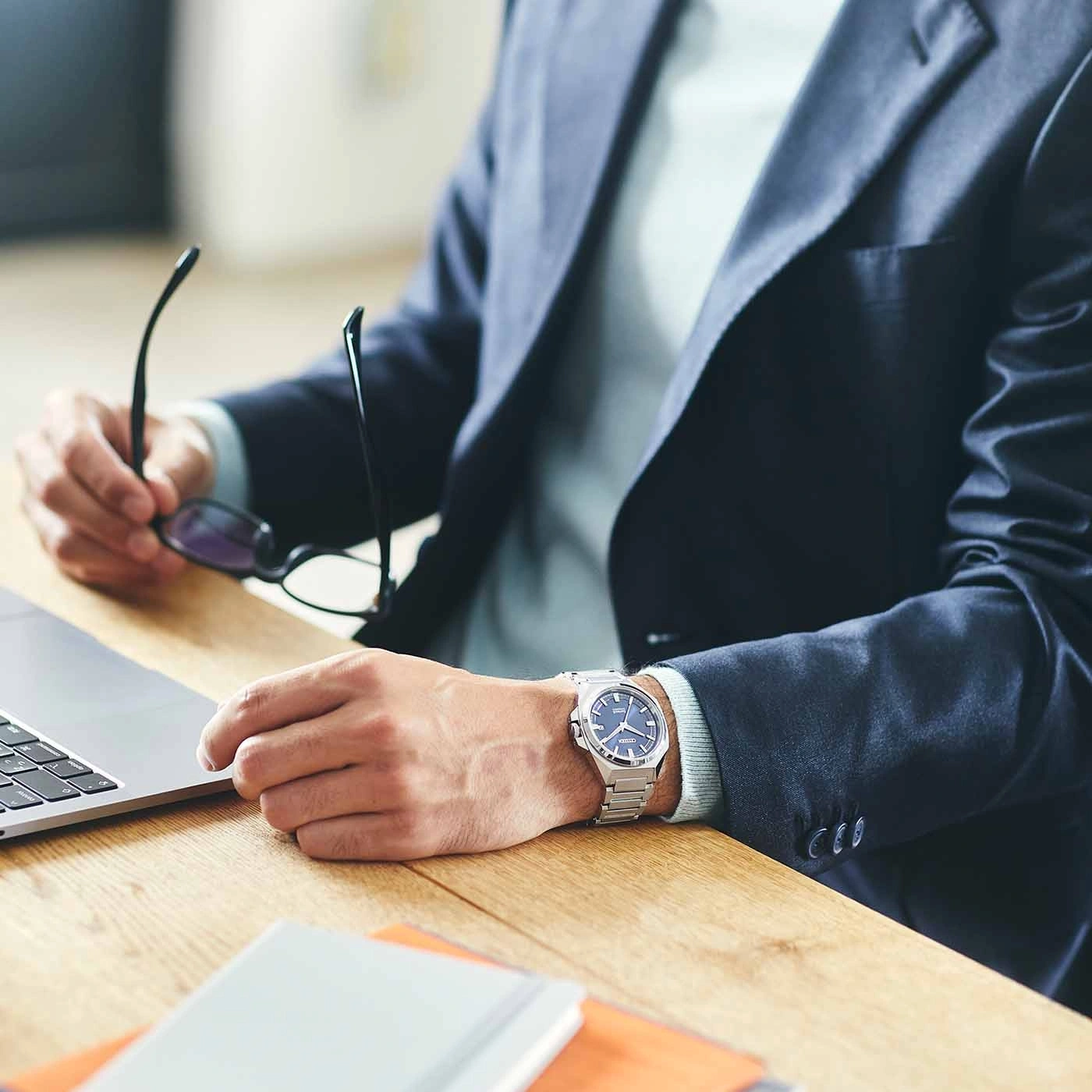 Man in suit with watch, holding glasses, at a desk with a laptop.
