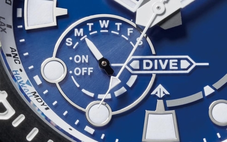 A close-up shot of a blue watch face featuring day of the week indicators (M, T, W, T, F, S) and an "ON/OFF" switch, alongside other details like "DIVED" and city abbreviations.
