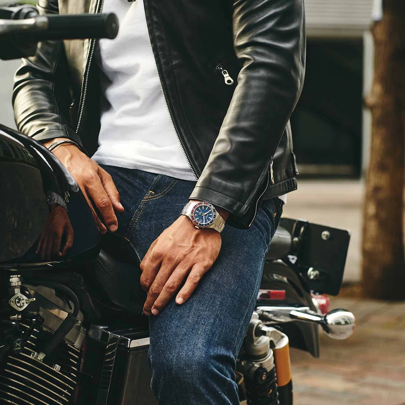 Man on motorcycle wearing watch and leather jacket, jeans.