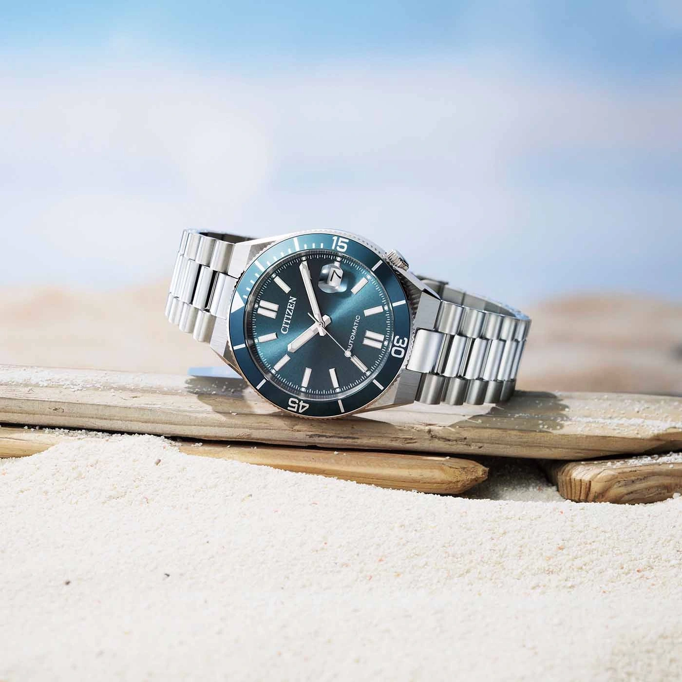 A Citizen automatic watch rests on driftwood on a sandy beach.