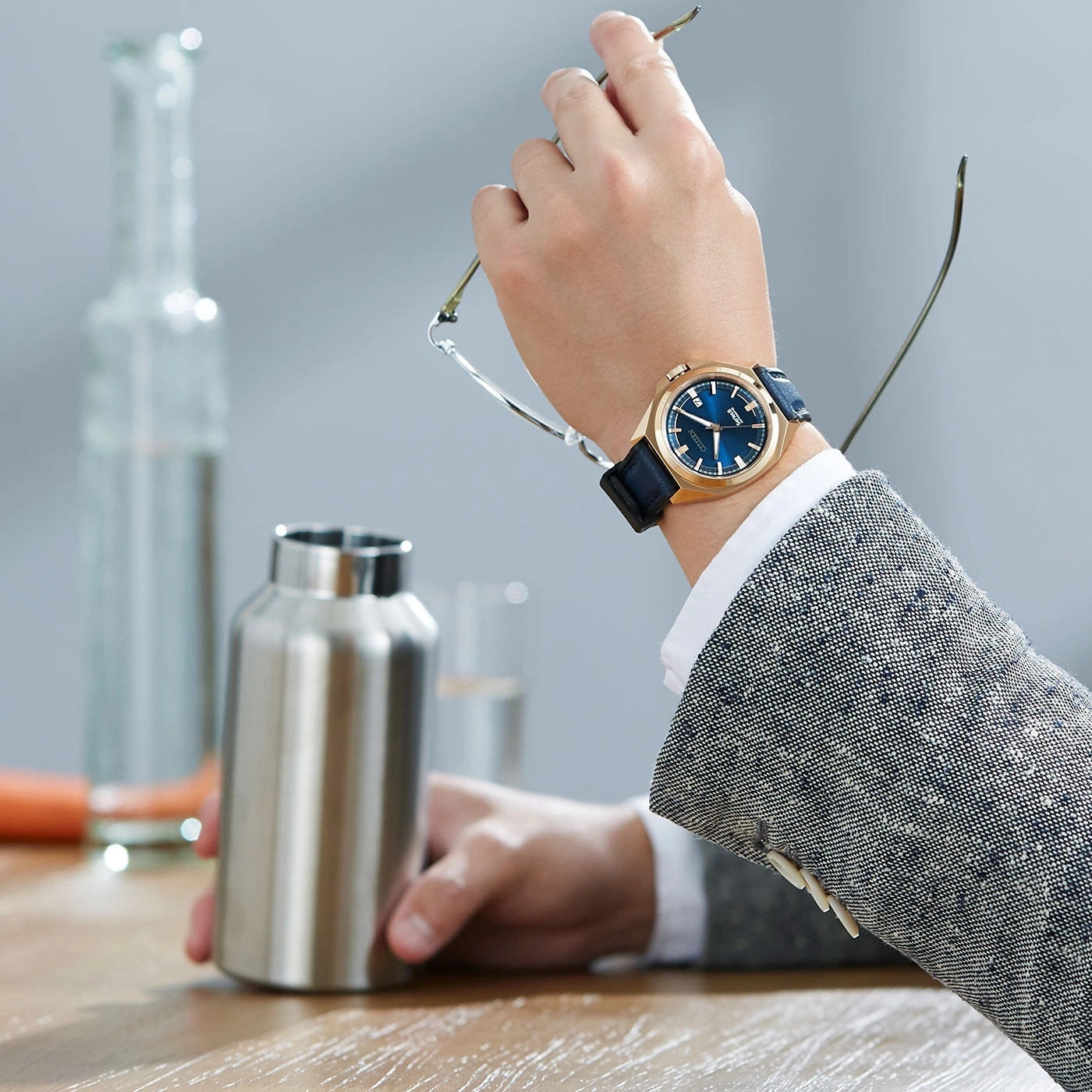 Man wearing a watch, holding glasses, with a water bottle on a table.