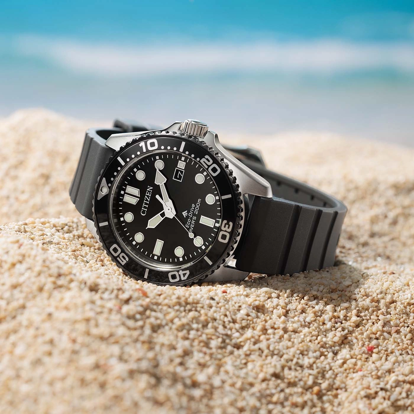 A black Citizen Eco-Drive dive watch sits on sandy beach with ocean background.