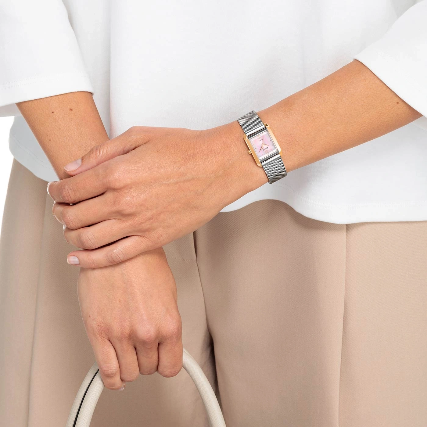 Woman wearing a watch, holding a bag, wearing white shirt and beige pants.