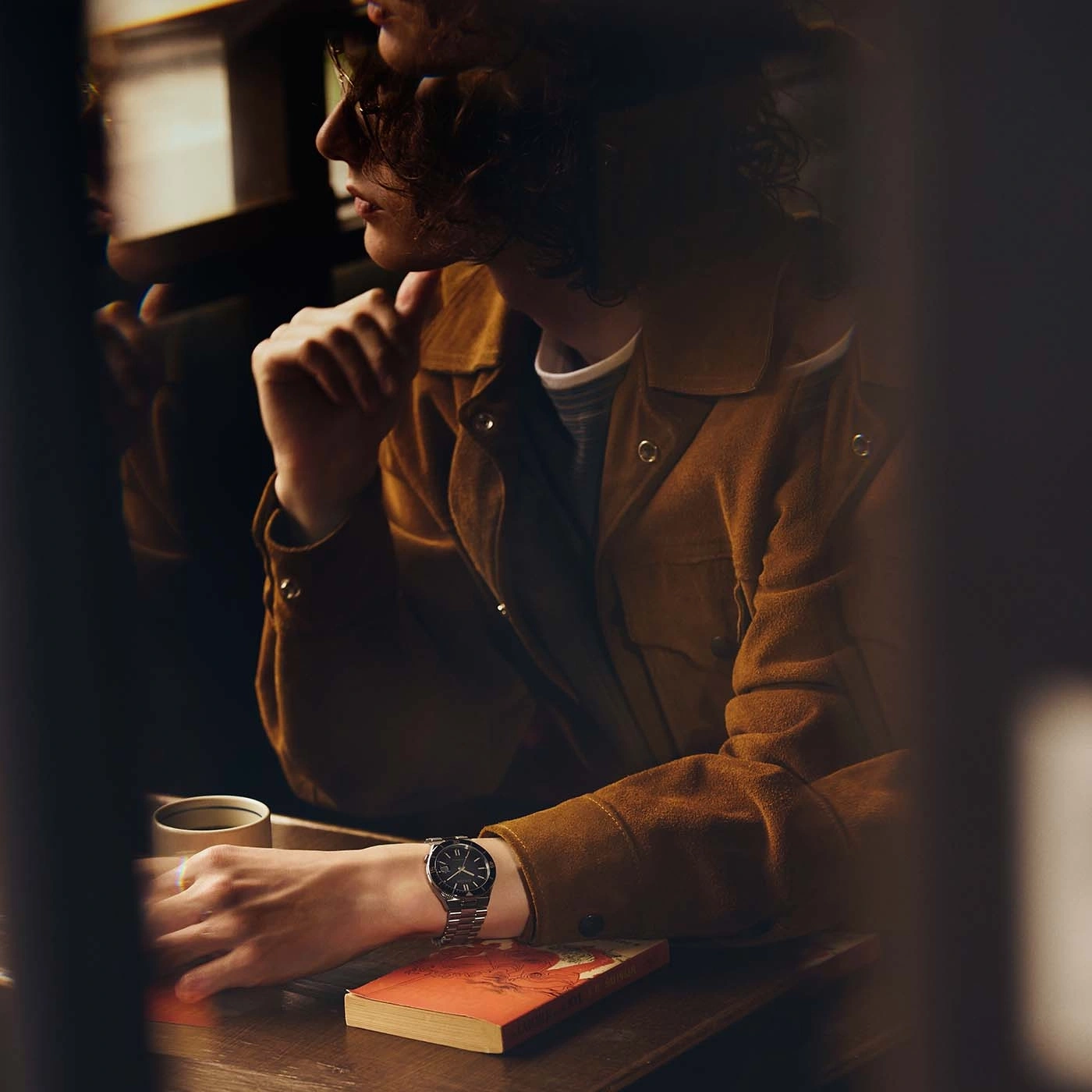 Close-up of a person wearing a watch and jacket, sitting at a table with a book.