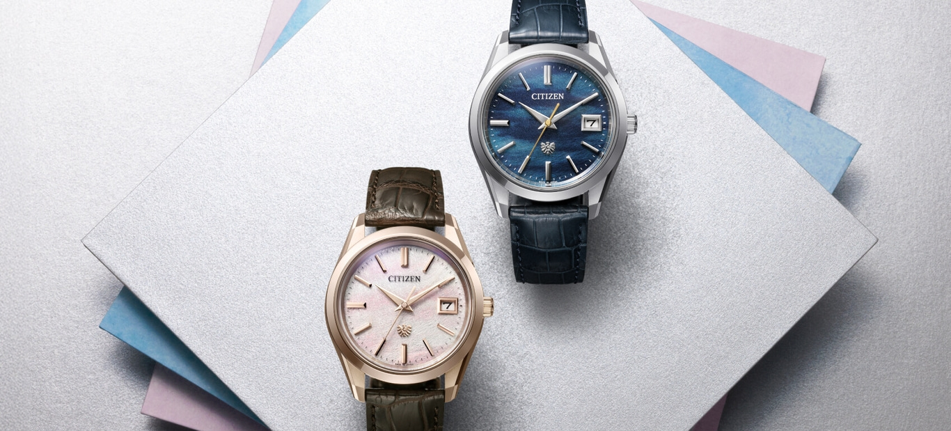 Two Citizen watches on a silver surface with colorful paper accents.