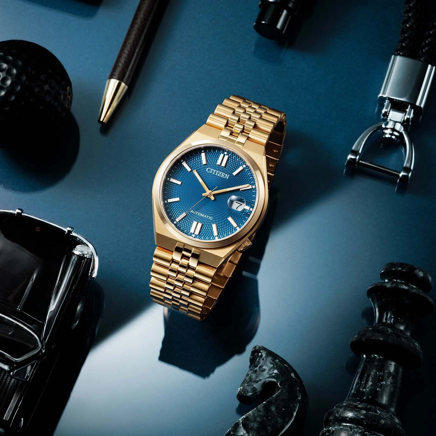 Gold watch with blue face, surrounded by accessories on blue surface.