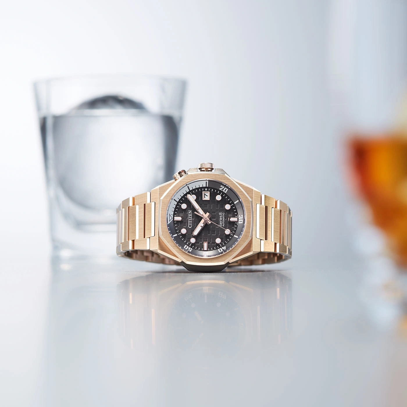 Gold watch with black face, glass of water, and reflection on surface.