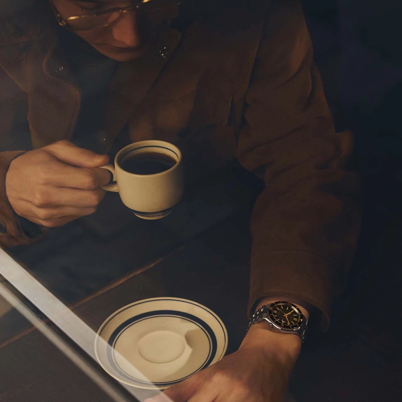 Man in brown jacket drinks coffee, wearing a watch.